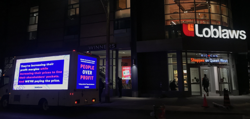 A billboard van parked outside of a Loblaws on a darkened street. The billboard says People over profit.